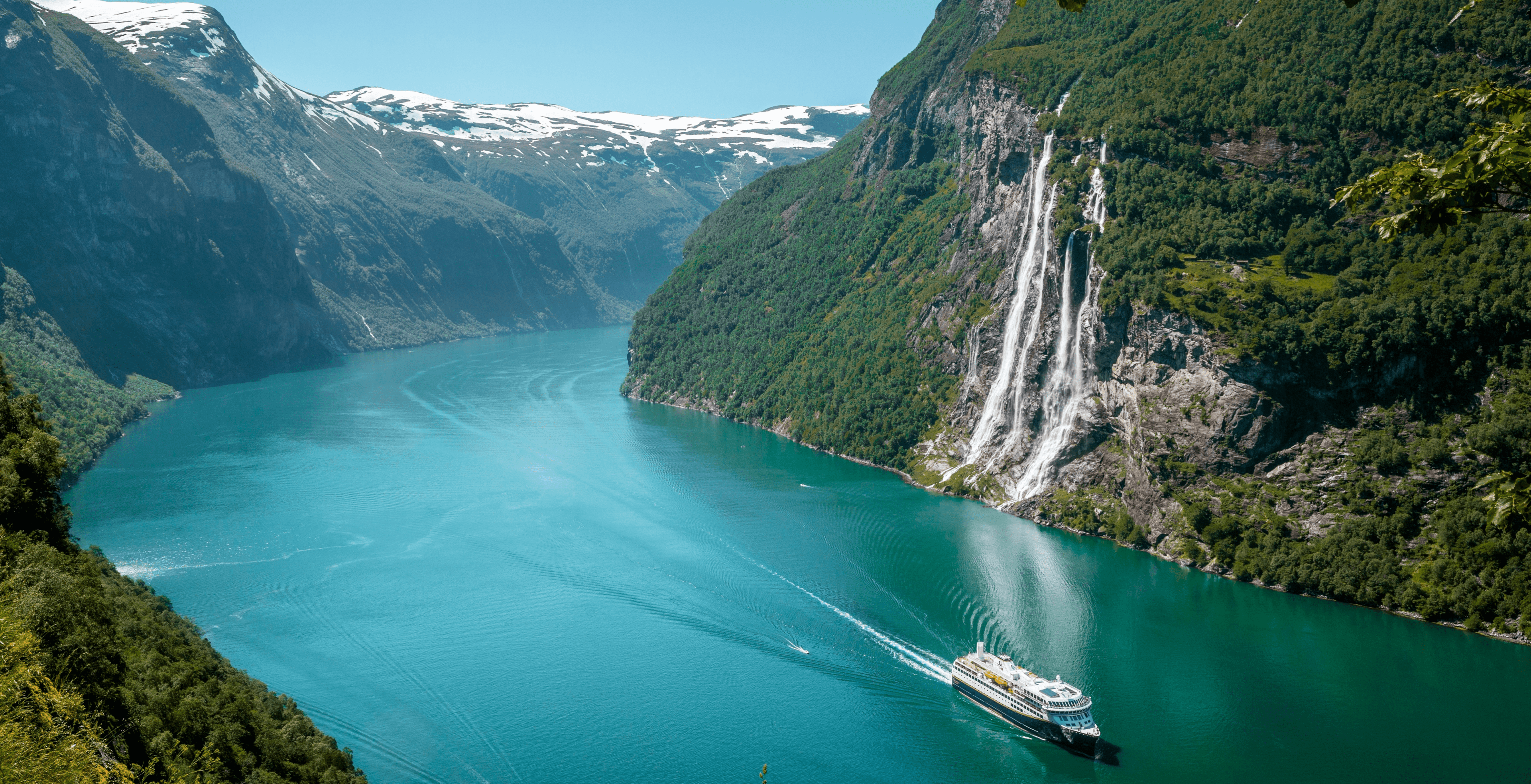 A distant shot of a cruise liner in a wide river gorge, flanked by majestic, snow capped, mountains on each side.