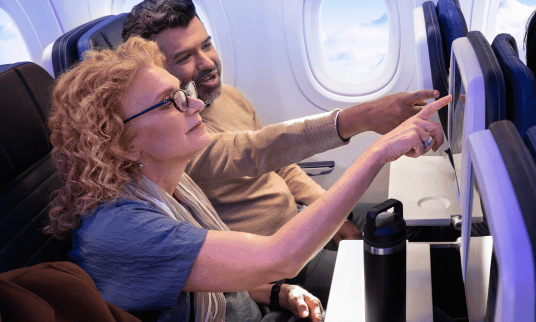 A man and woman on a plane both pointing at  a seatback display.