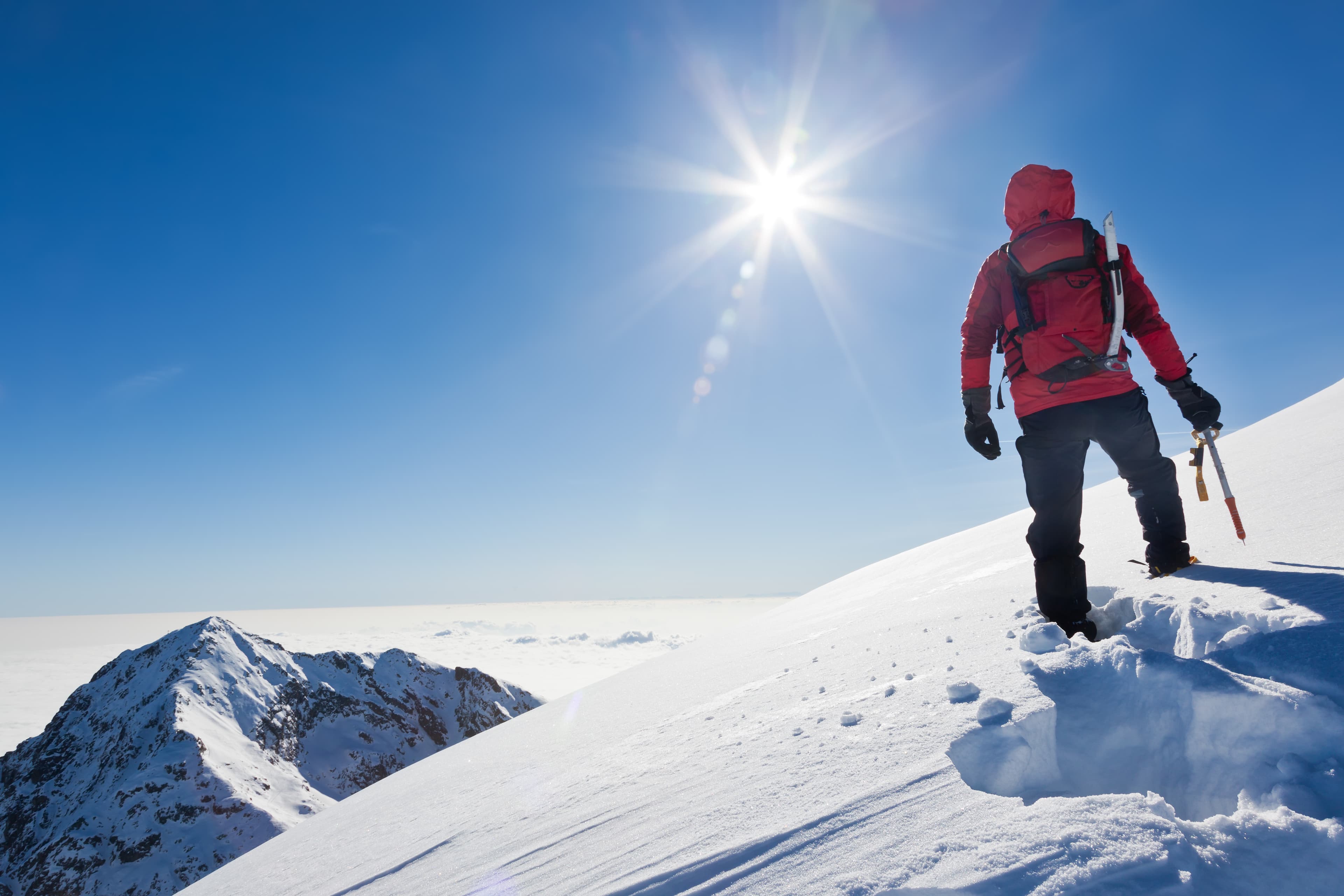 A mountain climber with a red jacket and climbing gear standing atop a snow covered mountain with a distant snow covered mountain peak to his left
