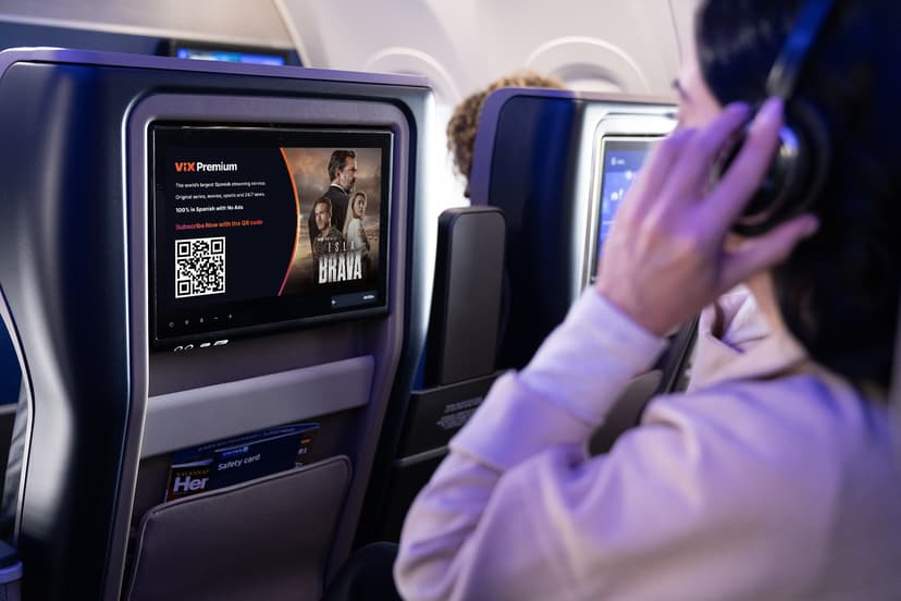 Woman sitting on a plane, watching the seat back monitor while she touches her wireless headphones.
