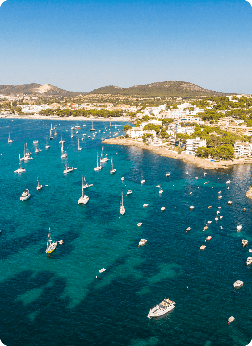 Aerial view of a coastal town with white buildings along the shore and numerous boats anchored in turquoise blue waters